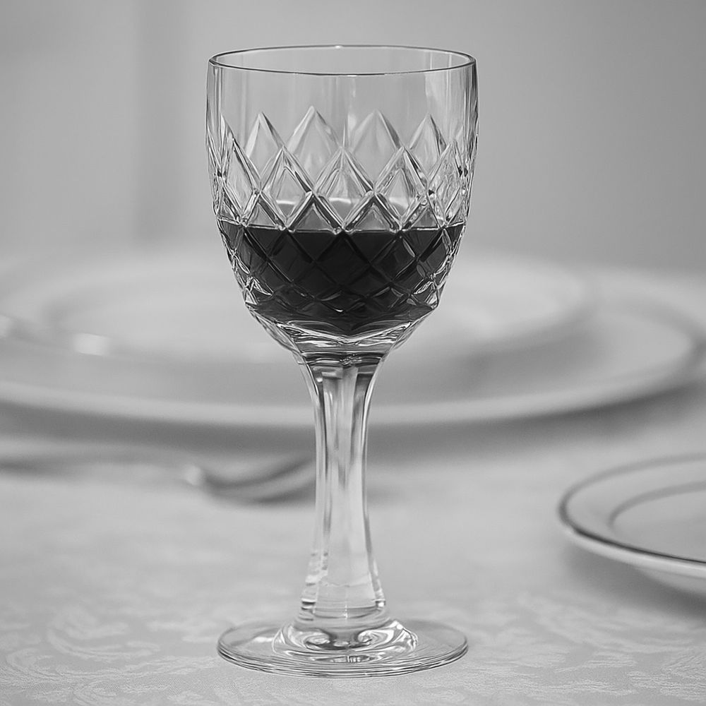 Ein elegantes Kristall-Weinglas vom Typ Venedig mit Diamantschliff steht auf einem stilvoll gedeckten Tisch. Es ist halb mit dunklem Rotwein gefüllt und reflektiert das Licht. Im unscharfen Hintergrund: weiße Teller.